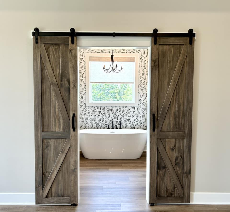 Rustic sliding barn doors open to a bathroom with a freestanding tub, leafy wallpaper, chandelier, and window.