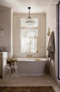 Freestanding white soaking tub beneath a tall window with sheer curtains, gold fixtures, and a chandelier in a bright marble bathroom.