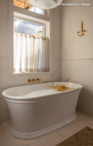 White freestanding bathtub with gold fixtures in a marble bathroom, window with sheer curtains, and a bath tray.