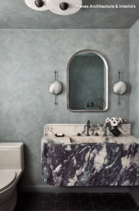 Blue-green textured bathroom featuring a marble sink, arched mirror, and two round wall sconces.
