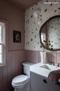 Pink-walled bathroom with wainscoting, floral wallpaper, round wall-mounted mirror, and a white sink with a vase of flowers above the vanity.