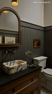 Bathroom with dark beadboard walls, marble vessel sink on a wooden vanity, and brass fixtures beside a white toilet.