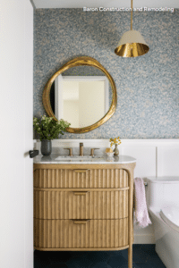 Bathroom with a curved wooden vanity, round gold mirror, blue patterned wallpaper, and a white toilet nearby (decor-focused).