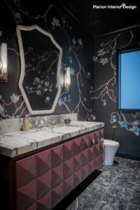 Bathroom interior with marble-topped vanity and geometric burgundy cabinets, an octagonal mirror, floral wallpaper, and a toilet.