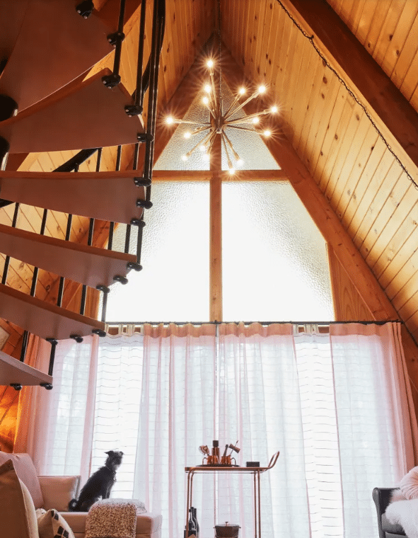 Cozy loft living room with a high wooden ceiling, spiral staircase, and a starburst chandelier; cat sits on an ottoman near sheer curtains.
