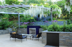From Patio to Paradise: How to Build Your Dream Garden Oasis Outdoor patio with a modern built-in fireplace, black metal seating, and a pergola draped with cascading white flowers over stone planter walls.