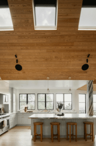 Beyond Just “Natural Light”: The New Engineering Standard for Home Skylights - Intrepid Builders LLC. Wood-paneled vaulted kitchen ceiling with skylights overlooking a white kitchen island and wooden stools below a row of windows.