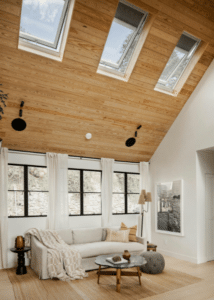 Beyond Just “Natural Light”: The New Engineering Standard for Home Skylights - Intrepid Builders LLC. Bright living room with a vaulted wooden ceiling and skylights, a white sofa with throw blankets, and a round coffee table.