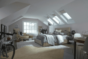 Beyond Just “Natural Light”: The New Engineering Standard for Home Skylights - Intrepid Builders LLC. Cozy attic bedroom with a low bed, gray bedding, and multiple skylights in a sloped ceiling room.