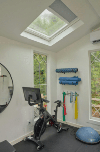 Beyond Just “Natural Light”: The New Engineering Standard for Home Skylights - Intrepid Builders LLC. Home gym with a stationary bike, wall-mounted foam rollers, resistance bands, and a blue exercise ball/bosu in a bright room with windows and skylight
