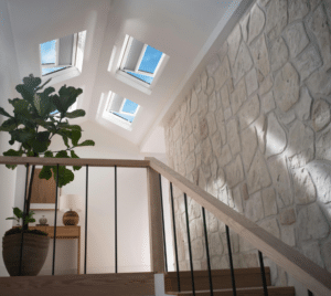 Beyond Just “Natural Light”: The New Engineering Standard for Home Skylights - Intrepid Builders LLC. Interior staircase with a stone wall, wooden railing, and a large potted plant reaching toward skylight windows. One wall features exposed stone texture and natural light streams in from above.