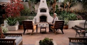 From Patio to Paradise: How to Build Your Dream Garden Oasis Older man sitting in wooden patio chairs facing a stone outdoor fireplace in a sunny courtyard with potted plants, creating a cozy backyard seating area.