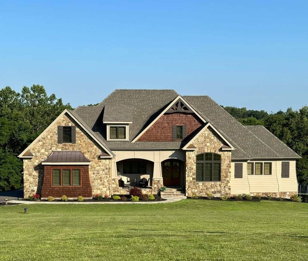 Intrepid Blog Front view of a large stone-and-siding house with a gabled roof and covered porch, set on a green lawn under a blue sky.