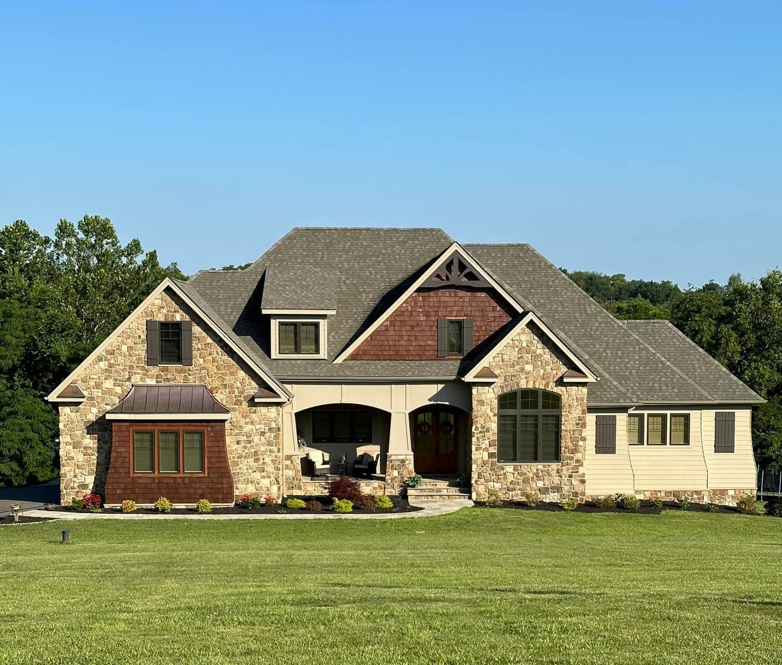 Front view of a large stone-and-siding house with a gabled roof and covered porch, set on a green lawn under a blue sky.